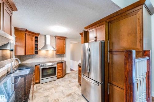 Kitchen featuring rich wood cabinetry, stainless steel appliances, dark stone countertops, tile backsplash, and tile flooring - 5212 142 Street Nw, Edmonton, AB - Indoor Photo Showing Kitchen With Double Sink