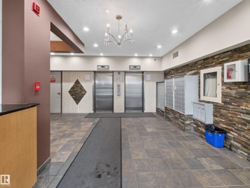 Lobby area featuring dual elevators with stainless steel doors, a stone veneer accent wall, and a chandelier with glass elements - #502 9710 105 St Nw, Edmonton, AB 
