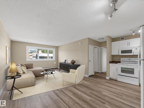 Open-concept living area featuring wood-finish flooring, white kitchen cabinetry, and large window - #502 9710 105 St Nw, Edmonton, AB 