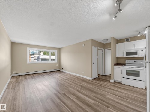 Open-concept living space featuring wood-finish flooring, a large window, white baseboard trim, and a kitchen area with white cabinetry, dark countertops, and white appliances - #502 9710 105 St Nw, Edmonton, AB 
