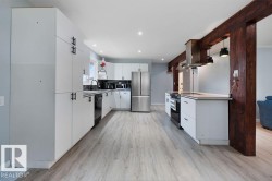 Modern kitchen featuring extensive white cabinetry with matte black hardware, stainless steel appliances, light wood-finish flooring, and a central island with a built-in range and overhead stainless steel range hood - 