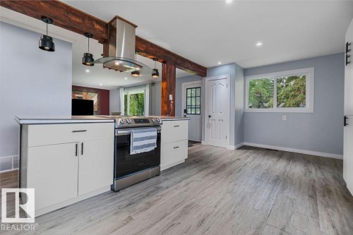 Kitchen featuring exposed wood beams, stainless steel range with overhead ventilation, light-toned wood-finish flooring, white cabinetry with dark hardware, and recessed lighting - 27 Apache Crescent, Leduc, AB 