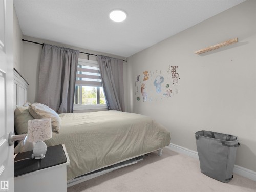 Carpeted bedroom featuring a window with blinds and drapes, a white paneled headboard, a ceiling light fixture, and a light-colored wall with baseboards - #30 4137 76 St Nw, Edmonton, AB 