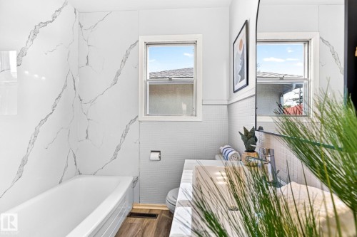 Contemporary bathroom featuring a tub with large format marble-look wall tiling, wood-finish flooring, a white vanity with an integrated sink, and dual windows - 16152 110B Avenue Nw, Edmonton, AB - Indoor Photo Showing Bathroom