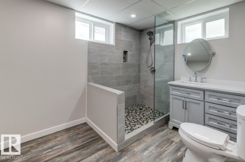Contemporary bathroom featuring a glass-enclosed shower with subway tile surround and pebble flooring, a light gray vanity with a white countertop and oval mirror, and wood-finish flooring - 8511 64 Avenue, Edmonton, AB - Indoor Photo Showing Bathroom