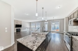 Open-concept kitchen and living area featuring a central island with a double basin sink, stainless steel appliances, wood-finish flooring, and a stone-clad fireplace - 