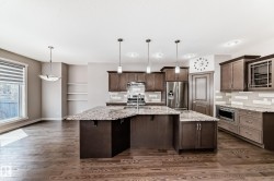 Spacious kitchen featuring a large granite-topped island with integrated sink, dark wood cabinetry, stainless steel appliances, and wood-finish flooring - 