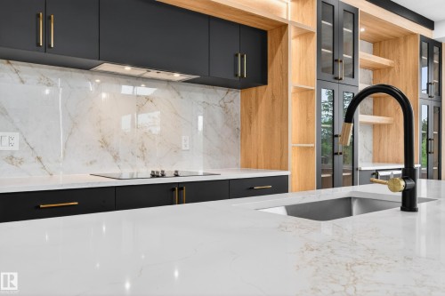 Kitchen featuring a white countertop island with an undermount sink and black and gold-tone faucet, black cabinetry with gold-tone hardware, wood-finish open shelving, white backsplash with veining, and a built-in stovetop - 431 172 Avenue, Edmonton, AB - 