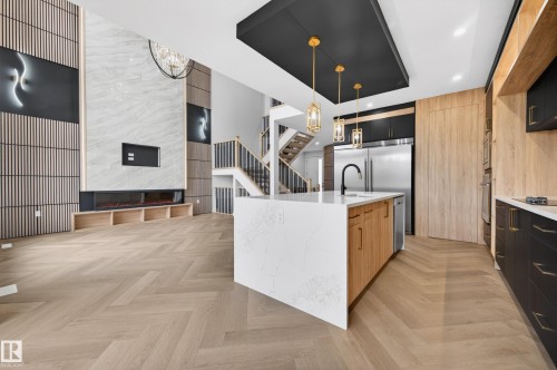 Open-concept living space featuring herringbone wood-finish flooring, a contemporary kitchen island with a white countertop and wood-finish cabinetry, and a striking fireplace wall with marble-finish paneling and wood slat accents - 431 172 Avenue, Edmonton, AB - Indoor Photo Showing Kitchen