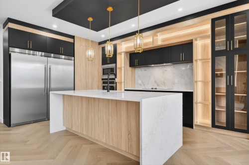 Modern kitchen featuring a large center island with a white countertop and wood-finish paneling, herringbone wood-finish flooring, stainless steel built-in appliances, black cabinetry with gold-tone hardware, and integrated lighting within open shelving - 431 172 Avenue, Edmonton, AB - Indoor Photo Showing Kitchen With Upgraded Kitchen