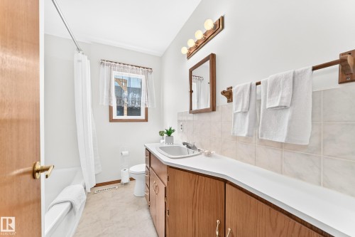 Bathroom featuring a white bathtub, a single window, a wood-finish vanity with a white countertop, a wall-mounted mirror, and tile wainscoting - 10819 137 Avenue Nw, Edmonton, AB - Indoor Photo Showing Bathroom