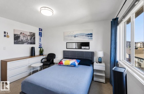 Light-filled bedroom featuring a large window with white framing and blue curtain panels, a built-in flush-mount ceiling light, and neutral carpet flooring - 17924 61 Street Nw, Edmonton, AB - Indoor Photo Showing Bedroom