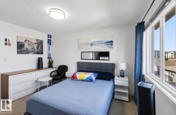 Light-filled bedroom featuring a large window with white framing and blue curtain panels, a built-in flush-mount ceiling light, and neutral carpet flooring - 