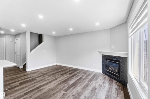 Living area featuring wood-finish flooring, recessed lighting, and a corner fireplace with a dark stone surround and white mantel - #64 120 Magrath Rd Nw, Edmonton, AB - Indoor With Fireplace