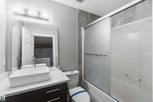 Modern bathroom featuring a white vessel sink on a light-colored countertop, a dark wood-finish vanity with drawer pulls, and a bathtub with sliding frosted glass doors - #64 120 Magrath Rd Nw, Edmonton, AB - Indoor Photo Showing Bathroom