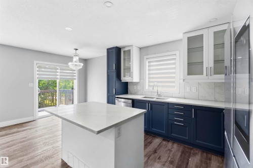 Contemporary kitchen featuring a central island with a light-toned countertop, two-tone cabinetry with dark lower and white upper units, a stainless steel sink with a modern faucet, and wood-finish flooring - #64 120 Magrath Rd Nw, Edmonton, AB - Indoor Photo Showing Kitchen