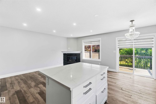 Kitchen island with white cabinetry and a light-toned countertop, wood-finish flooring, recessed lighting, and a fireplace with a dark stone surround and white mantle - #64 120 Magrath Rd Nw, Edmonton, AB - Indoor With Fireplace