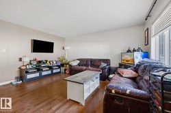Spacious living area featuring wood-finish flooring, neutral wall tones, a large window with vertical blinds, and a mounted flat-screen television - 