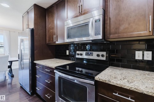 Kitchen featuring dark wood-finish cabinetry with brushed nickel hardware, light granite countertops, and a black subway tile backsplash - 17348 6 Avenue Nw, Edmonton, AB - Indoor Photo Showing Kitchen With Upgraded Kitchen