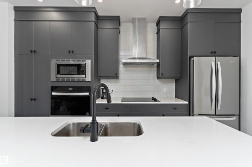 Sleek kitchen featuring gray cabinetry, stainless steel appliances, a double basin sink with a black faucet, white countertops, and a full-height tiled backsplash - 1439 Goodspeed Lane Nw, Edmonton, AB - Indoor Photo Showing Kitchen With Double Sink With Upgraded Kitchen