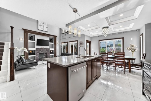 Open-concept living space featuring sleek tile flooring, a central kitchen island with integrated dishwasher, a built-in entertainment center with a stone-clad fireplace, and multiple coffered ceiling details with recessed lighting - 16244 138 Street Nw, Edmonton, AB - Indoor Photo Showing Kitchen With Double Sink