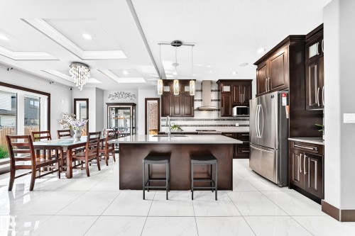 Spacious open-concept kitchen and dining area featuring white tile flooring, dark wood cabinetry, a large kitchen island with counter seating, stainless steel appliances, and a tray ceiling with recessed lighting - 16244 138 Street Nw, Edmonton, AB - Indoor