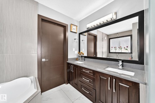 Contemporary bathroom featuring a large black-framed vanity mirror, a double vanity with dark wood cabinetry and integrated sinks, a grey countertop, and a built-in tub with textured tile surround - 16244 138 Street Nw, Edmonton, AB - Indoor Photo Showing Bathroom