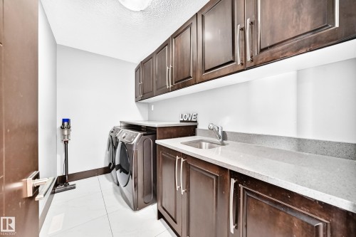Dedicated laundry area featuring dark wood-finish cabinetry, light-toned countertops, a stainless steel sink with a chrome faucet, and white tile flooring - 16244 138 Street Nw, Edmonton, AB - Indoor Photo Showing Laundry Room