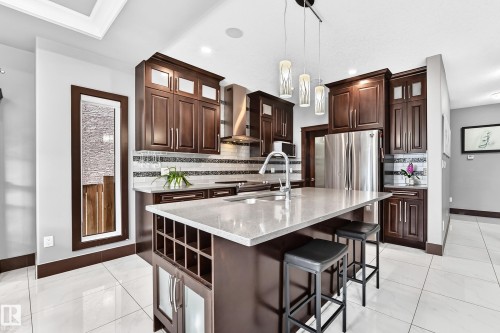 Gourmet kitchen featuring a central island with integrated wine storage, stainless steel appliances, dark wood cabinetry, and a sleek tile backsplash - 16244 138 Street Nw, Edmonton, AB - Indoor Photo Showing Kitchen With Upgraded Kitchen