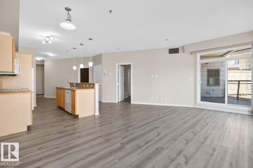 Open concept living area featuring wood-finish flooring, a kitchen island with sink, pendant lighting, and a sliding glass door to an outdoor area - #100 4835 104A St Nw, Edmonton, AB 