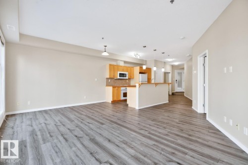 Open-concept living space featuring wood-finish flooring, a kitchen island with pendant lighting, light wood cabinetry, and stainless steel appliances - #100 4835 104A St Nw, Edmonton, AB 