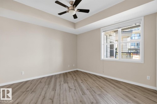 Spacious room featuring wood-finish flooring, a tray ceiling, and a ceiling fan with integrated lighting - #100 4835 104A St Nw, Edmonton, AB 