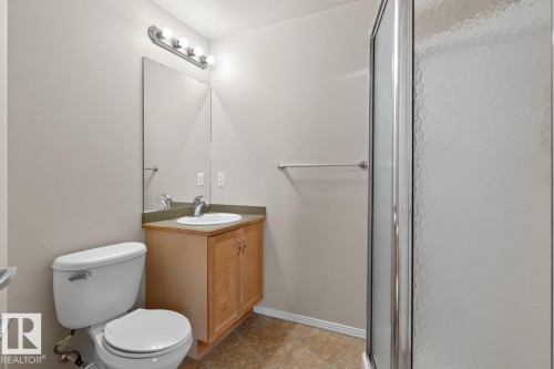 Bathroom featuring a light wood-finish vanity with a single basin sink, a large wall-mounted mirror, a four-bulb vanity light fixture, and a glass-enclosed shower with textured privacy glass - #100 4835 104A St Nw, Edmonton, AB 