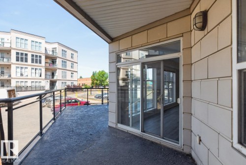 Exterior balcony featuring a textured dark surface, glass panel railing with a black metal frame, and a white sliding glass door - #100 4835 104A St Nw, Edmonton, AB 