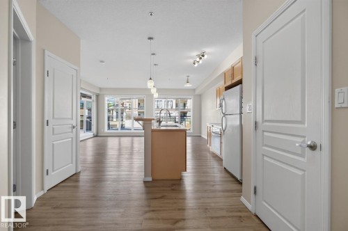 Open-concept living space featuring wood-finish flooring, a kitchen island with pendant lighting, ample natural light from large windows, light-toned cabinetry, and white appliances - #100 4835 104A St Nw, Edmonton, AB 