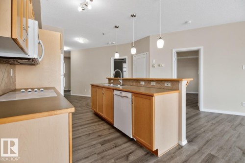 Kitchen featuring a central island with a double basin sink, light wood-finish cabinetry, and a white built-in dishwasher - #100 4835 104A St Nw, Edmonton, AB 