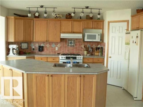 Kitchen featuring an island with double sinks, light wood-finish cabinetry, track lighting, a tiled backsplash, and light-colored countertops - 1686 Tompkins Wynd Nw, Edmonton, AB - Indoor Photo Showing Kitchen