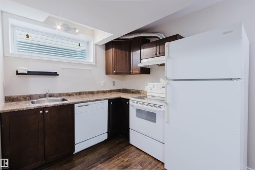 Kitchen featuring wood-finish flooring, dark wood cabinetry, laminate countertops, a stainless steel sink, and white appliances including a refrigerator, dishwasher, and electric range - 1686 Tompkins Wynd Nw, Edmonton, AB - Indoor Photo Showing Kitchen