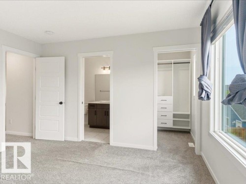 Carpeted interior room featuring a large window, a built-in closet system with shelving and drawers, and access to a separate bathroom with a vanity, mirror, and wall-mounted light fixture - 17487 77 Street, Edmonton, AB - Indoor