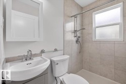 Three-piece bathroom featuring a shower with light-toned tile surround and a window, a white toilet, and a white vanity with a chrome faucet - 