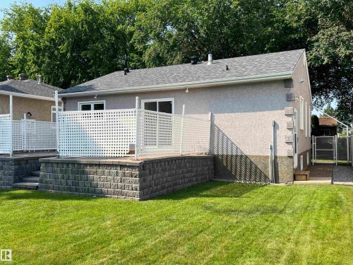 Expansive backyard featuring a manicured lawn, a raised patio with white lattice fencing, and a retaining wall constructed from stacked stone blocks - 9723 65 Avenue Nw, Edmonton, AB - Outdoor With Deck Patio Veranda With Exterior