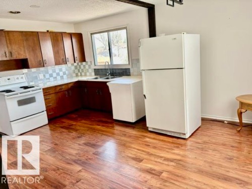 Kitchen featuring wood-finish flooring, wood cabinetry, white appliances, checkered tile backsplash, and a double basin sink - 5206 51Ave, Willingdon, AB - Indoor Photo Showing Kitchen With Double Sink
