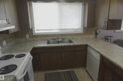 Kitchen featuring a double basin stainless steel sink, wood-finish cabinetry, tile backsplash, and light-toned countertops - 