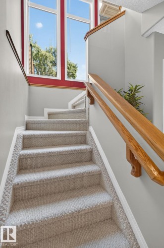 Carpeted staircase with light gray walls, wooden handrails, and a prominent window feature with red trim - 3211 Mcphadden Place Sw, Edmonton, AB - Indoor Photo Showing Other Room