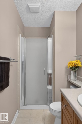 Compact bathroom featuring a shower enclosure with frosted glass panels, a light wood-finish vanity with a light-toned countertop, a toilet, and light-toned floor tiling - 3211 Mcphadden Place Sw, Edmonton, AB - Indoor Photo Showing Bathroom