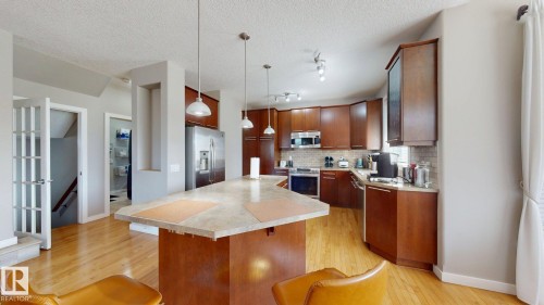 Spacious kitchen featuring an angular central island with pendant lighting, wood-finish flooring, cherry-toned cabinetry, stainless steel appliances, and a tiled backsplash - 335 62 Street Sw, Edmonton, AB - Indoor Photo Showing Kitchen With Upgraded Kitchen
