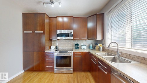 Modern kitchen featuring wood-finish cabinetry, stainless steel appliances, light-toned countertops, a subway tile backsplash, and wood-finish flooring - 335 62 Street Sw, Edmonton, AB - Indoor Photo Showing Kitchen With Double Sink
