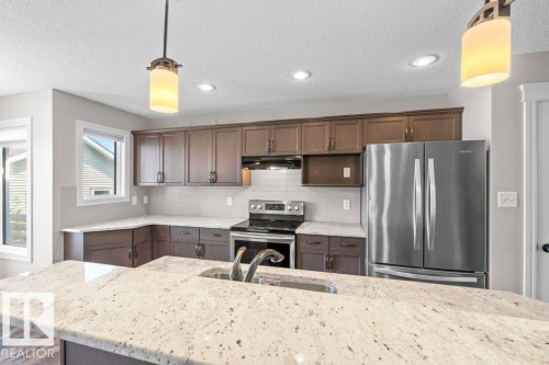 Spacious kitchen featuring a large island with an undermount sink, light-toned countertops, rich wood-finish cabinetry, stainless steel appliances, and recessed lighting - 1627 167 Street Sw, Edmonton, AB - Indoor Photo Showing Kitchen With Double Sink