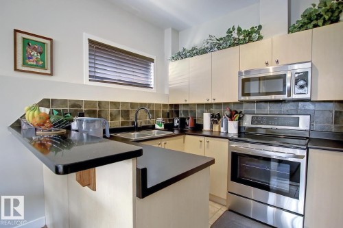 Compact kitchen featuring a black countertop peninsula, stainless steel appliances, and cream-colored cabinetry - 102 10905 109 Street, Edmonton, AB - Indoor Photo Showing Kitchen With Double Sink