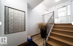 Interior common area featuring mailboxes, wood-finish flooring, a staircase with a black metal railing, and a glass-paneled entrance door - 